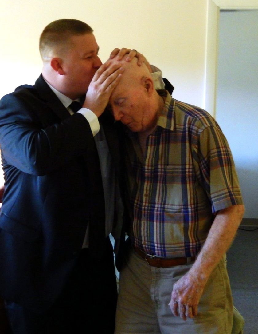 Luke Bernhardt in a dark suit kissing the forehead of his grandfather Alan Thevenet, holding his head in both hands